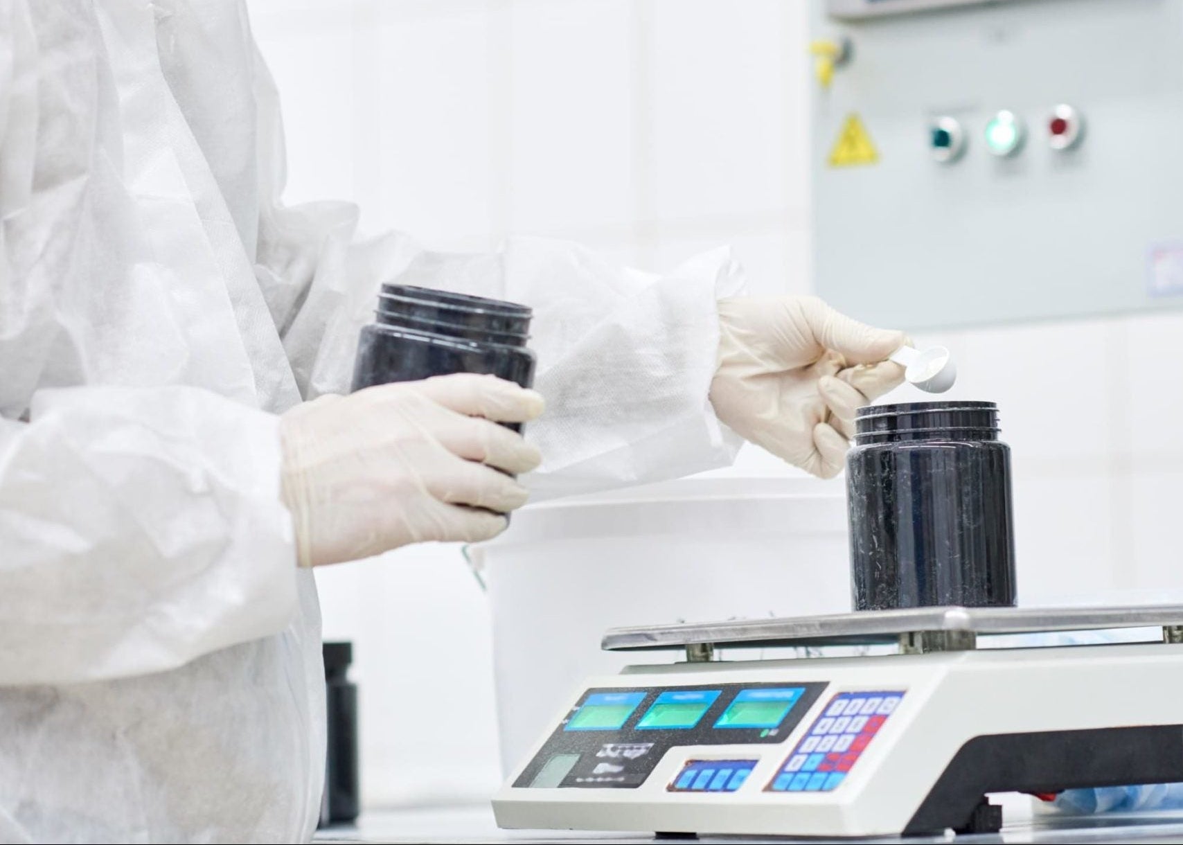 Person in a lab coat and gloves working with containers on a scale in a laboratory setting.