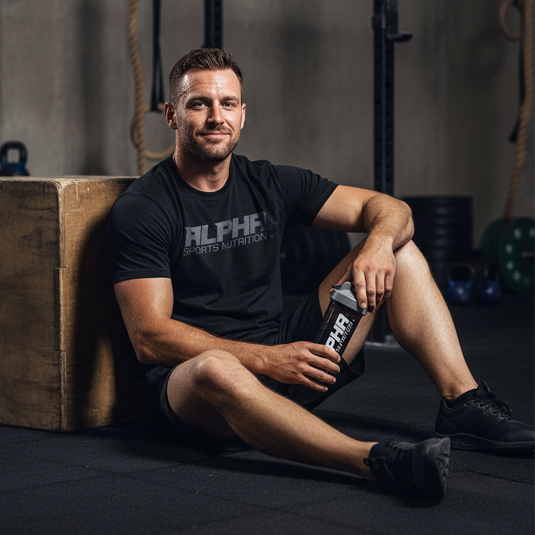 Man in a gym wearing an Alpha Sports Nutrition t-shirt, holding a supplement bottle.